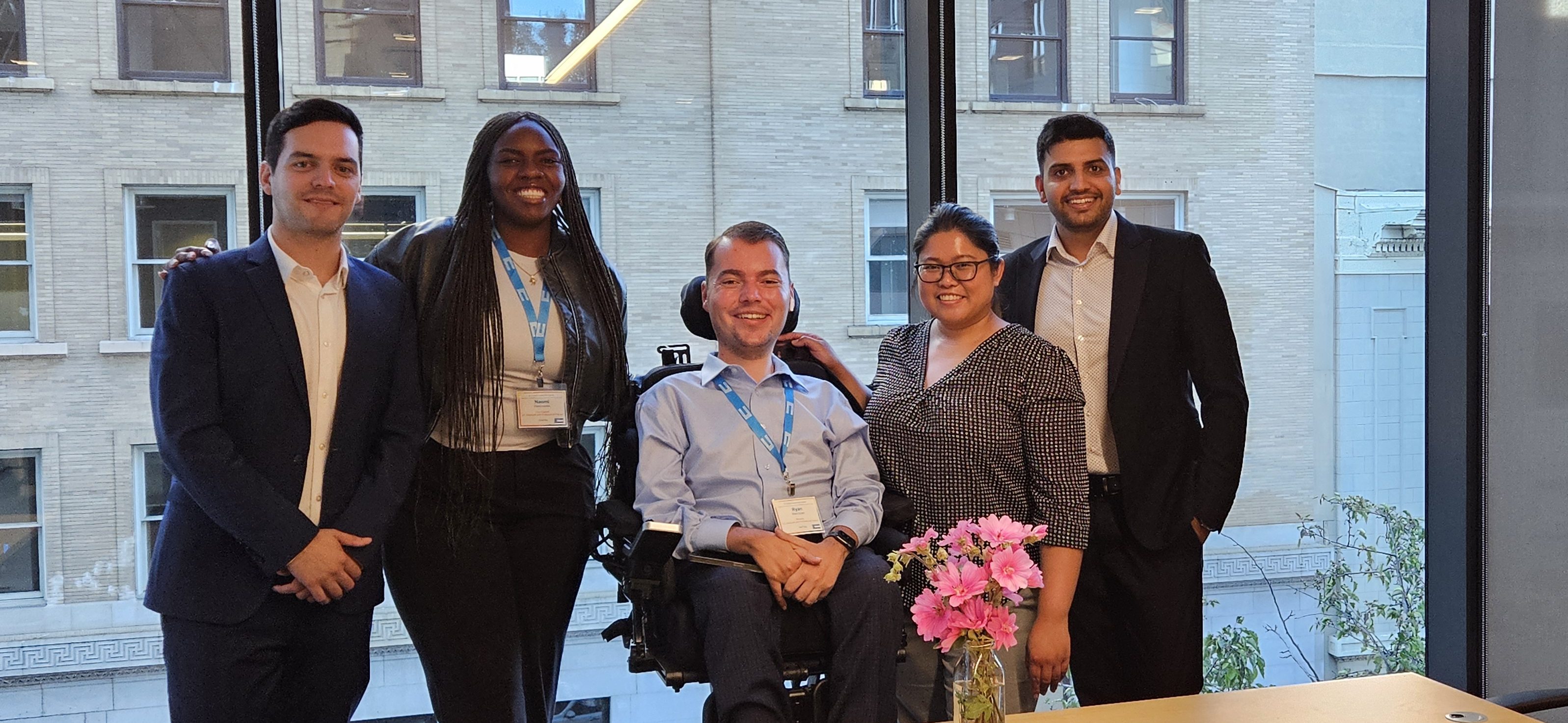 A group of five professional individuals posing together in an office setting with large windows, showcasing a vibrant city view. A floral arrangement is placed on the table in front of them.